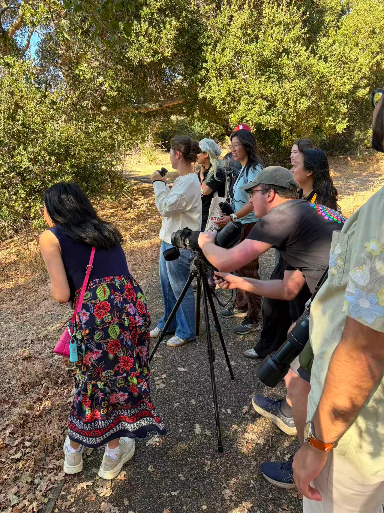 Members of the Stanford Birdwatching Club gathered on a tree-lined path with binoculars, cameras and a tripod-mounted spotting scope.